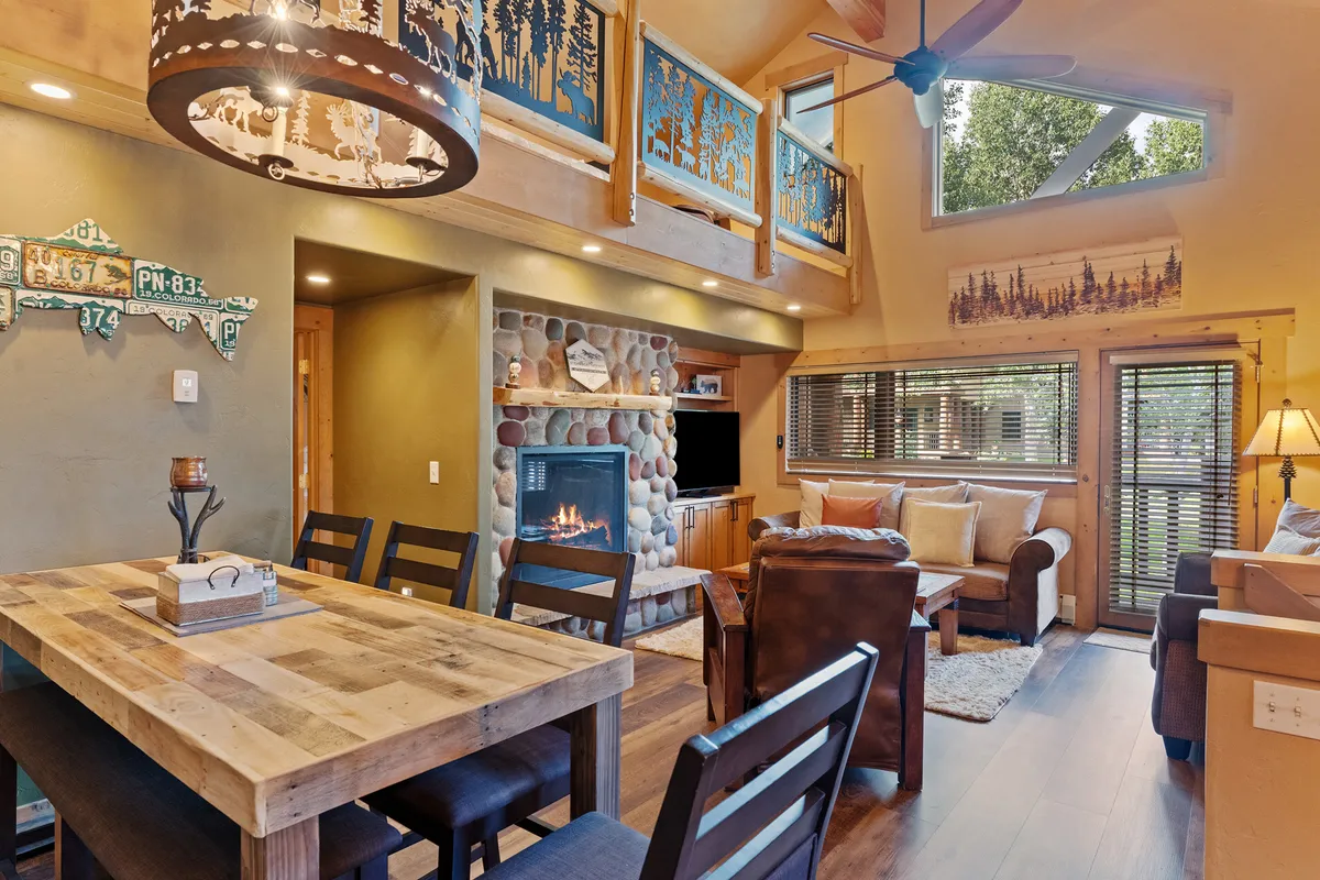 This image showcases a warmly lit, open-concept space featuring a striking wooden dining table positioned near a cozy gas fireplace. Large windows bathe the room in natural light, while a decorative railing and comfortable seating invite relaxation and group gatherings.