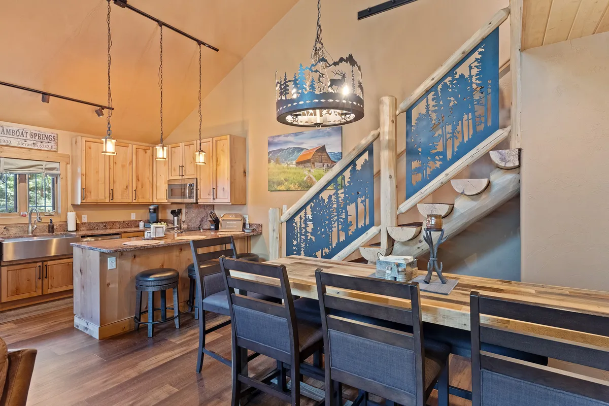 This inviting interior scene features a spacious kitchen island, an elegant dining table set for gatherings, a beautifully crafted staircase with an artistic railing, a striking chandelier, and a landscape painting that adds a touch of mountain charm. The space is bathed in warm light, creating a welcoming atmosphere ideal for cooking, dining, and entertaining after a day on the slopes.