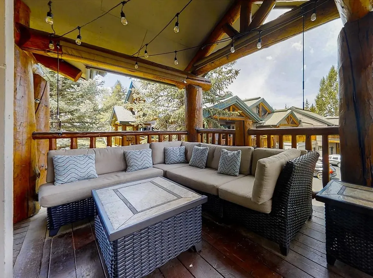 A cozy sectional sofa and wooden deck railing frame a welcoming outdoor lounge, complete with hanging string lights and natural surroundings. The scene suggests comfort and relaxation—ideal for gatherings, sipping morning coffee, or basking in sunset views.