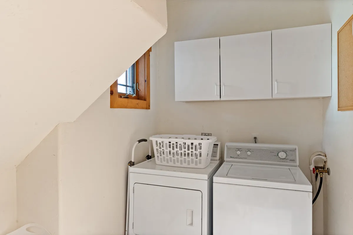 A well-organized laundry room featuring a washing machine, dryer, laundry basket, and upper storage cabinets. The room is clean, practical, and designed for efficient handling of laundry tasks.