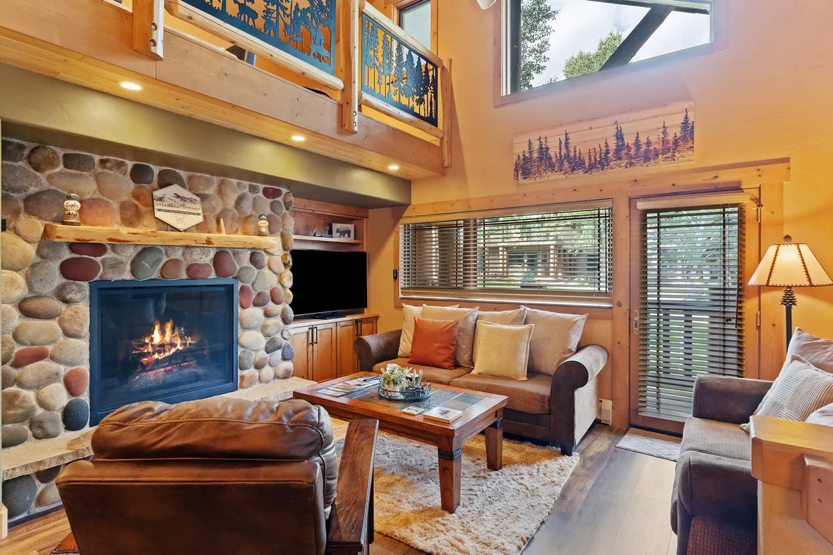 A welcoming living room anchored by a large sofa and coffee table, set near a fireplace and a wide window that floods the space with natural light. The arrangement encourages socializing, relaxing, or enjoying an evening by the fire.