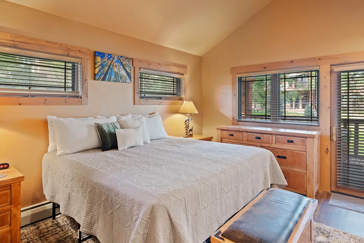 A serene bedroom featuring a well-made bed with neutral linens, a decorative painting above the headboard, a wooden dresser, and large windows that flood the room with natural light. The tranquil setup suggests a restful, private space ideal for unwinding.