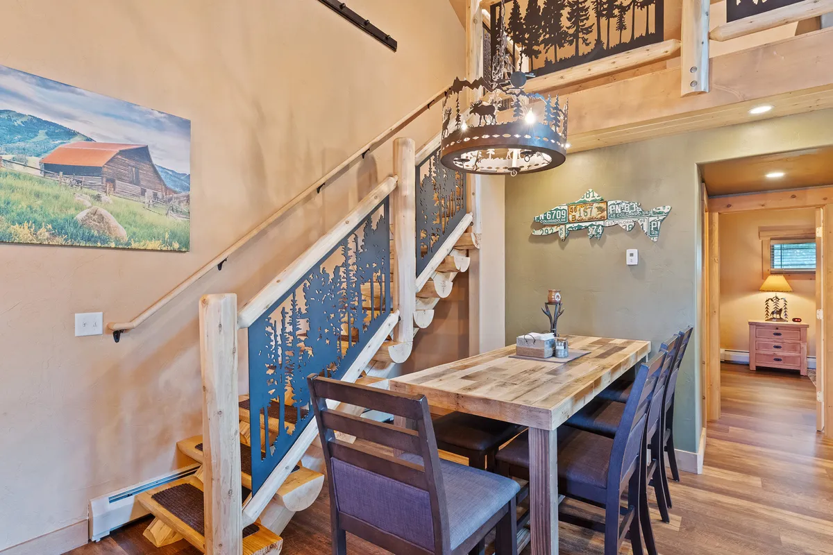 A spacious dining area with a wooden table under a striking metal chandelier. The scene suggests a place for family dinners, friendly gatherings, or casual meetings, set against a backdrop of an elegant staircase and framed landscape art.