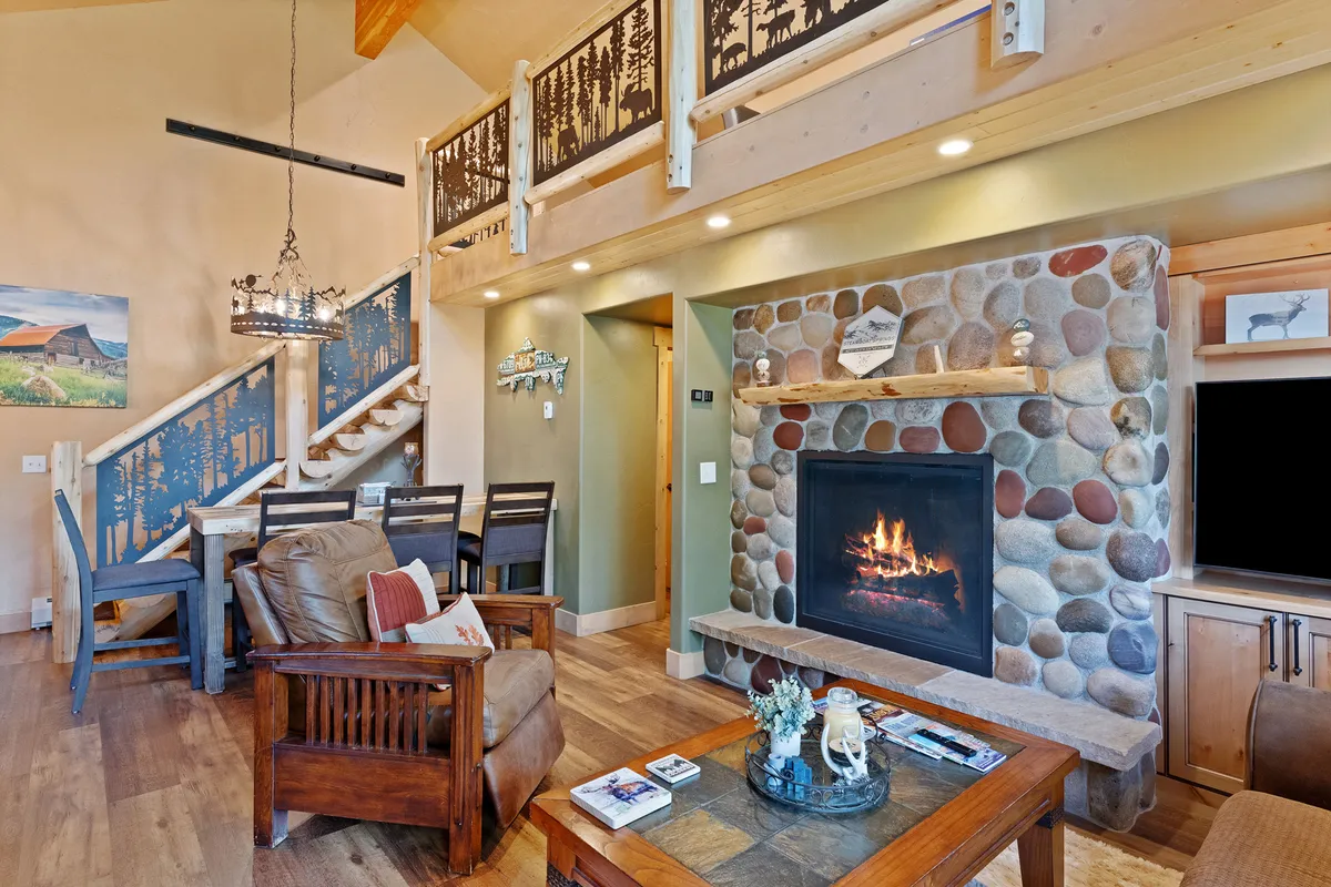 A spacious, warm living and dining area featuring a fireplace, wooden staircase, substantial dining table, and a cozy lounge chair. The space is set up for both group gatherings and intimate family moments, with local artwork adding a touch of character.