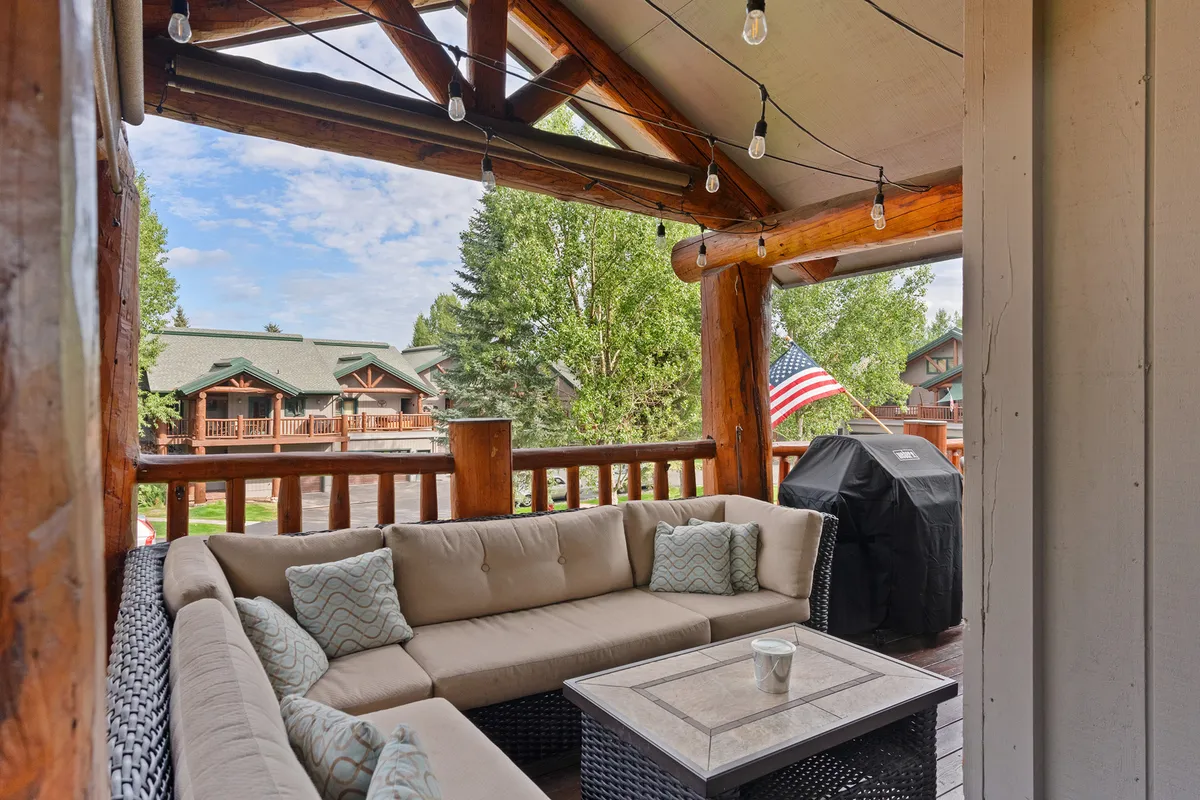 An exterior scene featuring a comfortable seating area on a deck, complete with a grill and framed by wooden beams. The space is set for relaxation and social gatherings, with hints of sunset views and string lights suggesting a seamless indoor-outdoor flow.